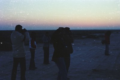 Silhouetted group of photographers at dusk, silhouetted against a soft sunset horizon. Vintage film grain overlay enhances no...