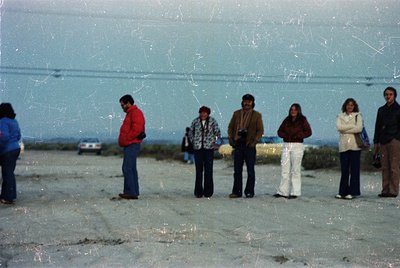 Vintage group photo on a concrete beachfront, likely 1970s–1980s. Six individuals in casual 70s attire—plaid shirts, bell-bot...