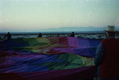 Vintage seaside scene featuring vibrant, hand-dyed fabric drying on a long rack. Four figures—two in foreground, two in backg...