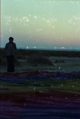 Vintage photograph of a lone figure silhouetted against a sunset, standing on a wet, reflective surface near water. The scene...