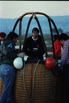 Hot-air balloon basket with woven rattan frame, carrying four individuals in casual 1970s attire—one holding a camera, anothe...