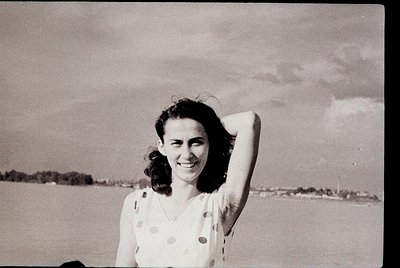 Vintage black-and-white portrait of a woman in a polka-dot dress, hand on head, smiling against a blurred seaside background....