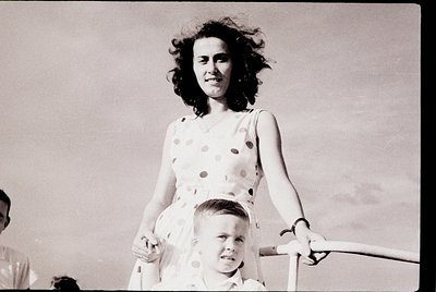 Black-and-white candid of a woman in a polka-dot dress (1950s–60s style) holding a young boy on a carousel horse, mid-ride. C...