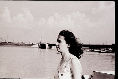 Black-and-white portrait of a woman in mid-20th century attire—polka-dotted blouse—standing near a waterfront with industrial...