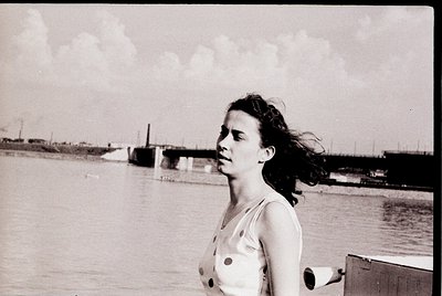 Black-and-white portrait of a woman in a sleeveless dress with button details, standing near a waterfront with industrial doc...