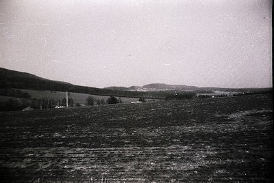 Vintage black-and-white landscape featuring rolling hills and sparse farmland. A single isolated house and scattered trees ma...
