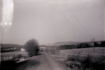 Vintage black-and-white rural road winding through open fields, bordered by sparse trees and utility poles. Overcast sky and ...