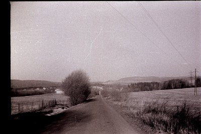 Vintage black-and-white rural road winding through open fields, bordered by sparse trees and fencing. Overcast sky and distan...