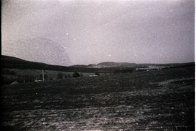 Vintage black-and-white landscape showing a rural horizon with sparse trees and distant buildings. Fog or mist obscures mid-g...