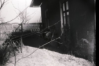 Vintage black-and-white photo of a rural porch with a child sitting on a makeshift swing made from branches. Wooden house wit...