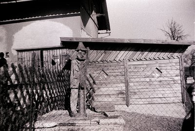 Black-and-white portrait of a man in a winter coat, hat, and gloves standing beside a rustic brick chimney and corrugated fen...