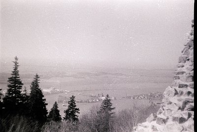 Black-and-white aerial view of a snow-dusted alpine valley, framed by rugged rock and coniferous trees. Distant village nestl...