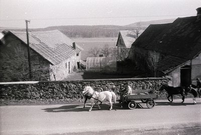 Mid-20th century rural scene: horse-drawn cart on unpaved road, guided by a person in a light jacket. Stone buildings with ti...