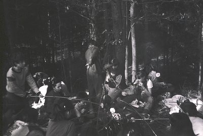 Group gathered around a campfire in a dense forest, mid-20th century. Wooden stumps and logs form makeshift seating; one pers...