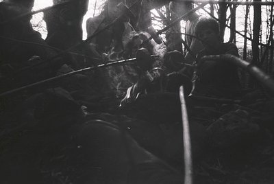 High-contrast black-and-white shot of two figures in dense forest, silhouetted against sunlight filtering through branches. O...