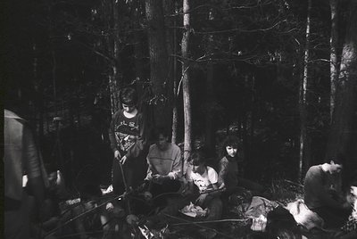 Four individuals gathered around a campfire in a dense forest, seated on logs and blankets. Mid-20th century outdoor lifestyl...