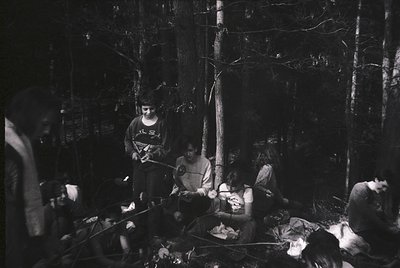 Black-and-white campfire scene in dense forest, likely 1970s–1980s. Group of 5–6 people seated on logs around a small fire, c...
