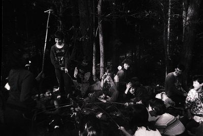 Black-and-white campfire scene in dense forest, likely 1970s. Group of 10+ people gathered around a fire pit, seated on logs....