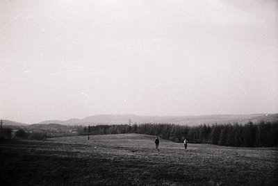 Black-and-white landscape featuring two figures walking across an open, slightly hilly field. Dense forest lines the horizon,...