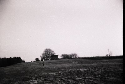 Vintage black-and-white rural landscape featuring a lone figure in mid-stride across an open field. Distant farmhouse and lea...