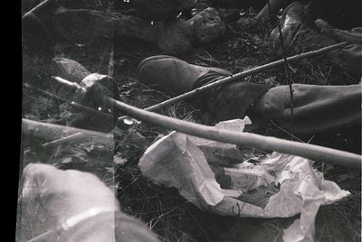 Black-and-white close-up of discarded items in a grassy area: crumpled paper, a broken bottle, and a twisted metal rod. Evide...