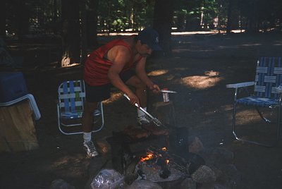 Man grills skewered meat over an open fire in a shaded outdoor picnic area. Foldable metal chairs and a cooler sit nearby. Wa...