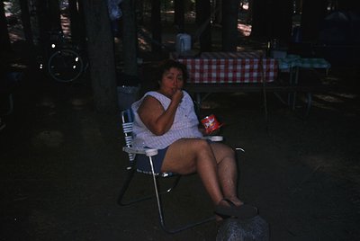 Vintage black-and-white photo of a woman seated on a metal chair in a dimly lit outdoor café, holding a small red container. ...