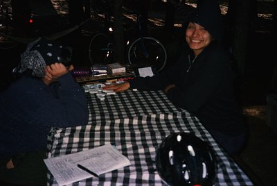 Vintage diner scene with two patrons seated at a checkered table, illuminated by warm overhead lights. One wears a hooded jac...