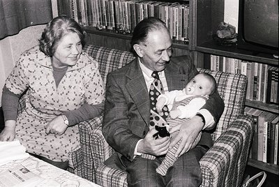 Mid-20th century (1950s-1960s) black-and-white family portrait: elderly couple in formal attire—man in suit & tie, woman in f...