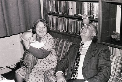 Mid-20th century black-and-white photo of two joyful adults in a cozy indoor setting. Woman in floral dress cradles a baby, m...