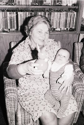 Mid-20th century indoor portrait: woman in floral dress holds baby in striped onesie, seated on armchair beside bookshelves. ...