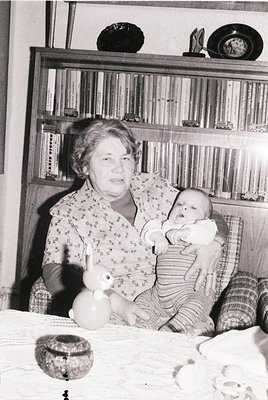 Mid-20th century indoor portrait of an elderly woman cradling a baby in a modestly furnished room. Wooden bookshelves filled ...