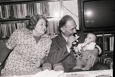 Black-and-white interior shot of elderly couple sharing a moment with a baby, likely mid-20th century. Man in suit holds baby...