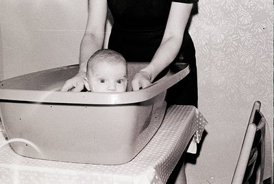 Mid-20th century indoor scene: infant in a metal baby tub, held by an unseen adult’s hands. Patterned wallpaper with floral m...