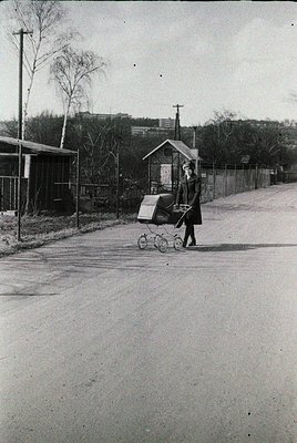 Mid-20th century street scene: Woman in long coat pushes a baby stroller with a white delivery bag on a paved road, flanked b...