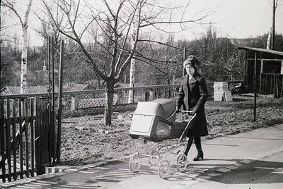 Black-and-white street scene featuring a woman in 1960s/70s fashion pushing a vintage pram with a basket on the handlebars. L...