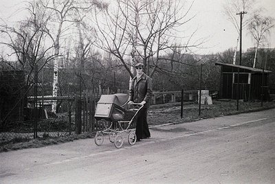 A woman in mid-20th century attire pushes a vintage pram along a quiet, tree-lined road. Her long coat and hat suggest a Euro...