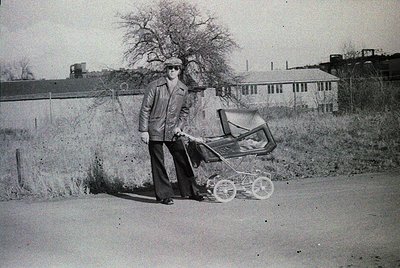 Man in 1970s-style wide-leg trousers and cap pushes a vintage pram on a rural road, surrounded by sparse trees and industrial...