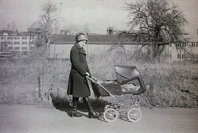 Mid-20th century woman in long coat and stockings pushes vintage pram with basket on urban sidewalk. Residential buildings an...