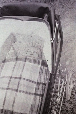 Vintage black-and-white photo of an infant wrapped in a plaid blanket inside a vintage stroller, likely mid-20th century. The...