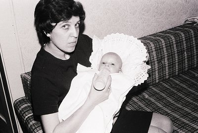 A woman in a short-sleeved blouse cradles an infant wrapped in a lace-trimmed bib, feeding them from a small glass bottle. Th...