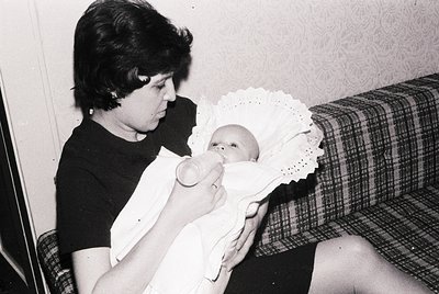 A woman feeds a swaddled infant in a mid-20th-century domestic setting. Floral wallpaper and a plaid couch suggest 1960s–1970...
