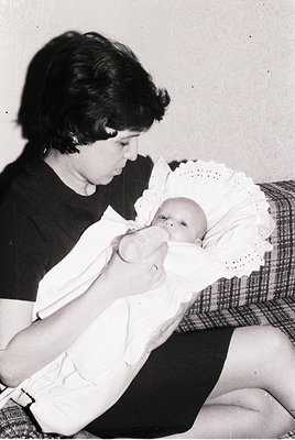 A tender 1950s-60s black-and-white photo of a woman nursing a swaddled infant in a plaid blanket, seated on a checkered couch...