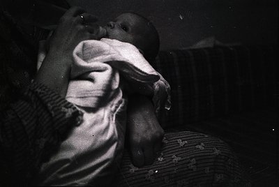 Black-and-white candid of an adult cradling an infant, likely mid-20th century. Soft focus and low lighting emphasize tender,...