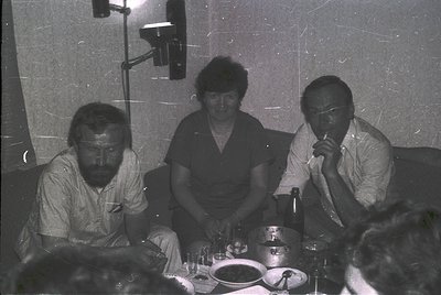 Three individuals seated at a table in a dimly lit studio setting, likely mid-1970s. The man on the left wears a striped butt...