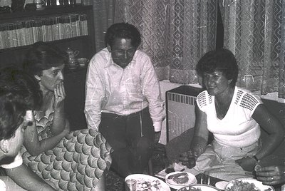 Black-and-white indoor gathering featuring four adults seated around a table laden with plates, glasses, and cutlery. Central...