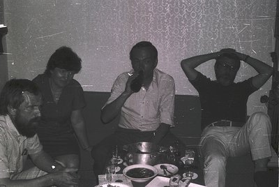 Vintage black-and-white photo of four adults seated indoors, likely mid-1970s. Casual setting with patterned wallpaper and a ...