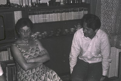 Two individuals seated indoors in a mid-20th-century setting, likely 1960s–1970s. Woman wears a patterned dress with geometri...