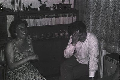 Vintage black-and-white indoor scene of two people laughing in a mid-century living room. Woman in a patterned dress sits lef...