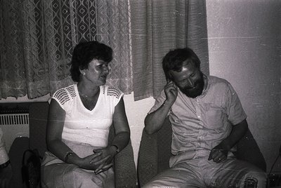 Two individuals seated indoors in a mid-20th-century setting, likely 1970s–1980s. Woman in a sleeveless blouse with geometric...